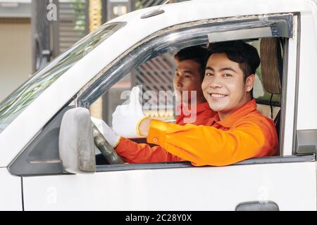 Homme asiatique portant une tenue orange assis en voiture montrant le pouce vers le haut tandis que son collègue conduit cette voiture, portrait horizontal Banque D'Images