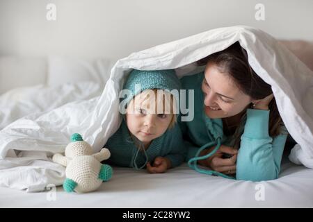 Bonne mère et enfant, garçon, avec une tenue assortie, allongé dans le lit, souriant heureux, embrassant et se cachant sous la couverture Banque D'Images