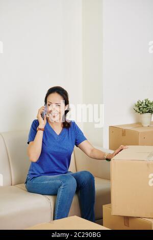 Une femme joyeuse assise sur un canapé après avoir été déménagent dans sa nouvelle maison, parlant au téléphone avec quelqu'un et touchant une boîte avec sa main Banque D'Images