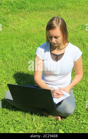 Une belle jeune fille blanche dans un t-shirt blanc et avec de longs cheveux assis sur l'herbe verte, sur la pelouse et travaillant derrière un ordinateur portable noir et écrit avec un stylo dans un bloc-notes. Banque D'Images