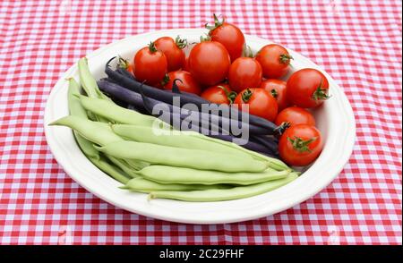 Yin Yang, haricots verts haricots verts violets et juteuses tomates cerises rouges dans un récipient blanc rond sur une nappe en vichy rouge Banque D'Images