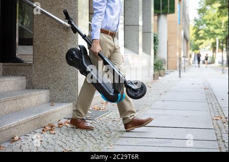 Homme portant l'E-scooter de travailler à la sortie de l'immeuble de bureaux Banque D'Images