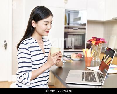 jeune femme d'affaires asiatique travaillant à la maison assise au comptoir de cuisine tenant une tasse de café regardant l'ordinateur portable Banque D'Images