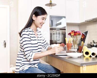 jeune femme d'affaires asiatique travaillant à la maison assis au comptoir de cuisine tenant une tasse de café regardant l'ordinateur portable, heureux et souriant Banque D'Images