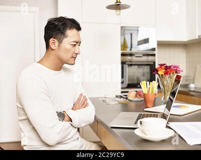 un jeune homme d'affaires asiatique travaillant à domicile assis au comptoir de cuisine et regardant les bras d'ordinateur portable a croisé Banque D'Images