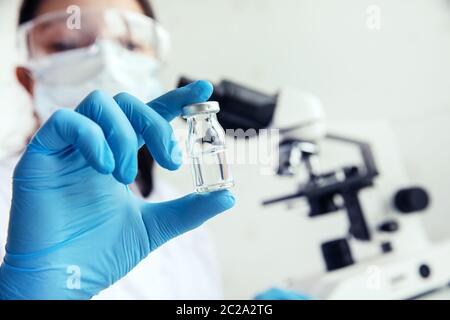 La main dans des gants médicaux tient le flacon. La médicienne tient le flacon dans sa main et un microscope dans le fond Banque D'Images