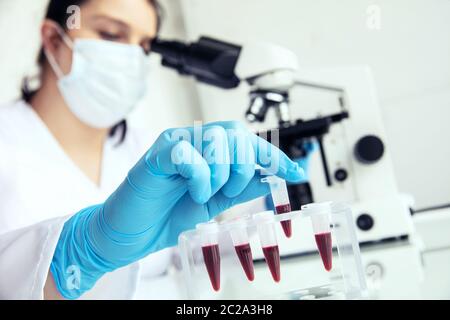 La main dans des gants médicaux prend eppendorf avec du sang. La médicienne femelle prend le tube à essai eppendorf avec du sang humain pour examiner et au microscope dans le backgro Banque D'Images