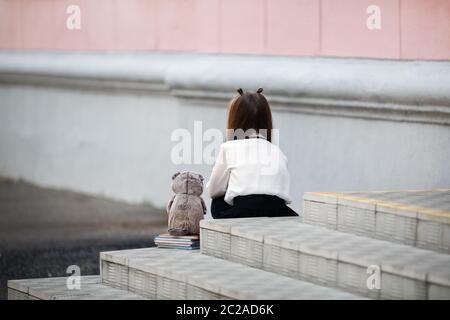 Triste fille avec une peluche est assis sur les marches de l'escalier de l'école avec son dos à la caméra. Enfant offensé. Focus sélectif. Banque D'Images