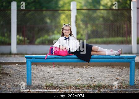 En uniforme d'écolière se reposant sur un banc. Smiling girl leaning on enfant sac à dos. Focus sélectif. Banque D'Images