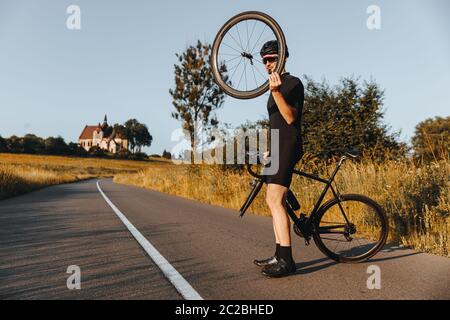 Homme mature en tenue active, casque de protection et lunettes miroir debout sur route pavée avec vélo cassé. Cycliste barbu tenant la roue à la main et Banque D'Images