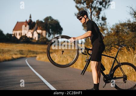 Vue latérale du cycliste barbu dans un casque de protection et des lunettes tenant une roue de son vélo tout en étant debout sur une route pavée. Concept de réparation et s Banque D'Images