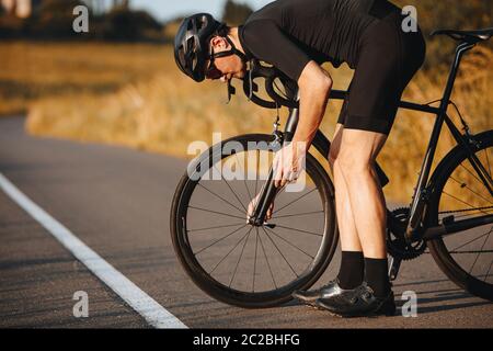 Vue latérale de l'homme actif dans des vêtements de sport se tenant courbé près de vélo noir pour fixer la roue. Sportif mature en casque noir prenant une pause pour réparer le vehi Banque D'Images