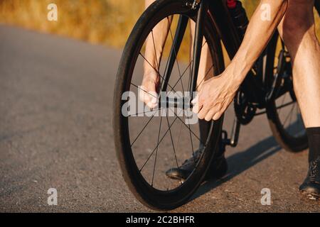 Gros plan d'un cycliste professionnel en mode activewear serrer les écrous sur la roue de vélo en extérieur. Homme fort répare le vélo sur route pavée. Banque D'Images