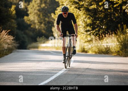 Sportif robuste avec corps sportif portant des vêtements de sport noirs, un casque et des lunettes de soleil s'exerçant en vélo à l'air frais. Concept de passe-temps sain a Banque D'Images