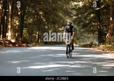 Portrait complet d'un sportif barbu en vêtements noirs, casque et lunettes de vue faisant de l'entraînement à vélo sur route pavée parmi la belle nature. Concept Banque D'Images