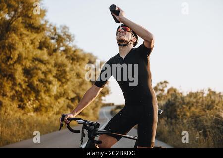 Homme mature fatigué en vêtements de sport, casque de protection et lunettes à miroir buvant de l'eau douce tout en se tenant sur une route pavée avec son vélo. Concept de Banque D'Images