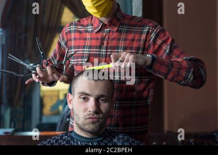 le coiffeur du coronavirus dans un masque protecteur coupe un jeune homme dans un salon de coiffure. Soins capillaires pendant la pandémie du coronavirus, Covid 19, coiffeur Banque D'Images