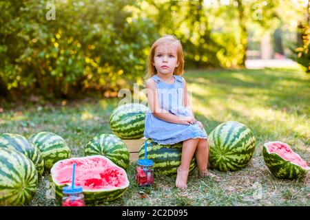 Jolie petite fille blonde assise avec des pastèques dans un parc. Été, fruits, concept de récolte Banque D'Images