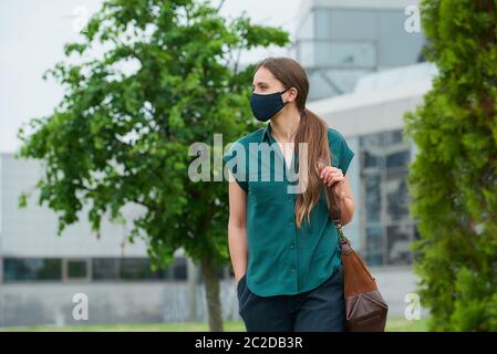 Femme dans le masque médical de visage marche près des arbres en portant une main dans la poche de pantalon, tient le sac dans le centre-ville. Fille gardant la distance sociale Banque D'Images