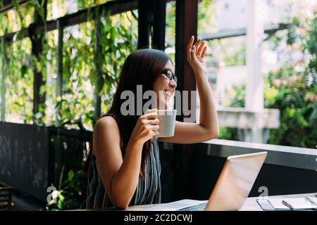 Jeune femme asiatique élégante souriant et agitant la main tout en étant assise à table avec un ordinateur portable et étudiant sur la terrasse Banque D'Images