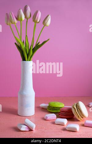 Les macarons et les petits guimauves blancs et roses sont dispersés sur un fond rose pâle à côté d'un vase de tulipes. Placer pour le texte Banque D'Images