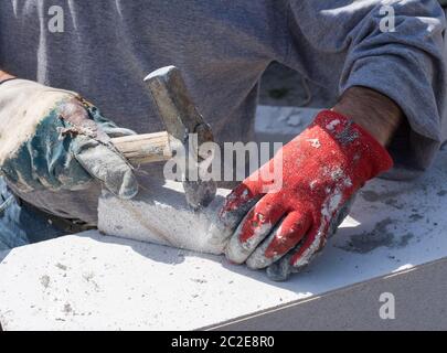 Travailleur avec des gants de travail vieux et sales frappant avec un bloc de béton blanc de marteau, de petits morceaux de béton volant dans l'air Banque D'Images