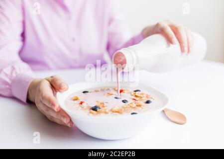 Gros plan de la femelle versant du yaourt aux fruits dans un bol avec des céréales, des fraises et des bleuets. Banque D'Images