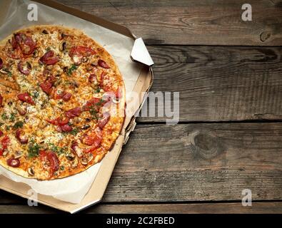 Ronde au four pizza avec saucisses fumées, champignons, tomates, fromage et de l'aneth dans une boîte en carton sur une table en bois, de l'alimentation est coupée en portions, t Banque D'Images