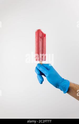 Main de l'homme tenant la glace rouge de la fraise popsicle, portant des gants bleus sur fond blanc. Prévention de la pandémie du coronavirus Banque D'Images