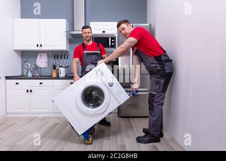 Deux jeunes hommes en uniforme des déménageurs professionnels de placer la machine à laver moderne dans la cuisine Banque D'Images