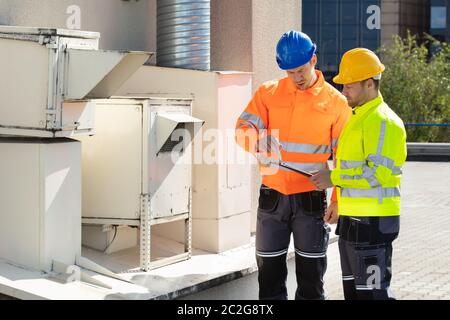 Deux électriciens hommes portant des vestes de sécurité Contrôle de l'unité de climatisation sur le toit du bâtiment Banque D'Images