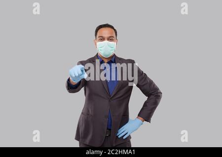 Homme d'affaires pointant devant regarder dans l'appareil photo portant un masque médical et des gants isolés. Homme d'affaires indien pointant du front Banque D'Images
