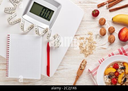 Balances blanches, ruban de mesure, carnet vierge, porridge de flocons d'avoine, baies, fruits et noix sur fond de bois clair. Vue de dessus. Concept de vies saines Banque D'Images