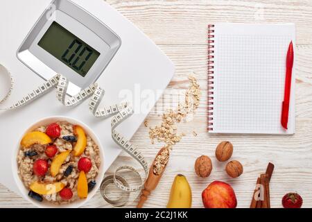 Balances blanches, ruban de mesure, carnet vierge, porridge de flocons d'avoine, baies, fruits et noix sur fond de bois clair. Vue de dessus. Concept de vies saines Banque D'Images