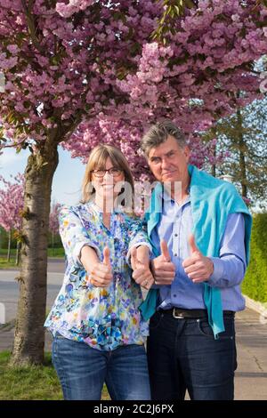 Un couple senior positif motivé qui se tient debout dans une rue bordée d'arbres contre un printemps rose, se fait un double pouce avec un sourire heureux Banque D'Images