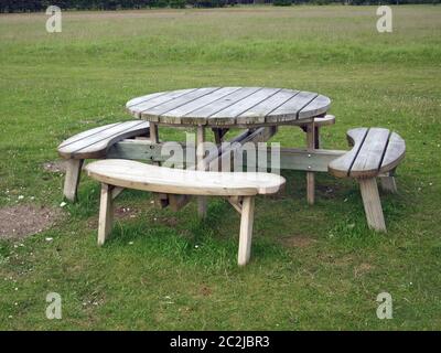 Table ronde de pique-nique en bois avec sièges arrondis sur un terrain d'herbe avec des arbres juste visibles en arrière-plan. Banque D'Images