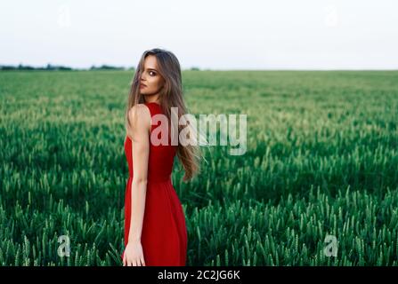 Belle jeune femme en robe rouge posant à l'extérieur dans le champ vert d'été Banque D'Images