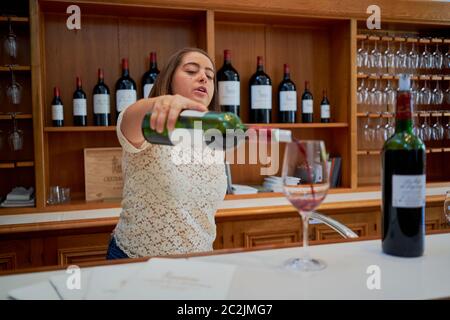 Une employée verse du vin rouge dans un verre au bar de dégustation du Château Lagrange, des bouteilles du domaine sur les étagères, Saint-Julien, France Banque D'Images