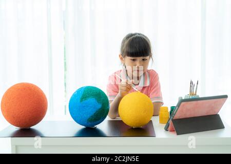 Une écolière asiatique élémentaire qui peint la lune en classe scientifique apprend sur le système solaire par vidéo conférence avec le professeur et d'autres camarades de classe Banque D'Images