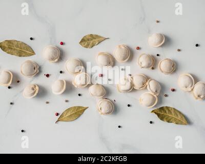 Schéma des pelmeni russes non cuits congelés avec des grains de poivre et les feuilles de laurier sur table en marbre blanc. Mise en page créative de boulettes. Belle dispersés ra Banque D'Images