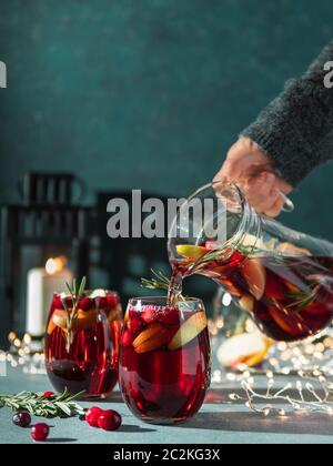 La main femelle verse la sangria d'hiver dans des verres avec tranche de fruits, canneberge et romarin. Arrière-plan sombre de noël avec bougie, décoration Lightti Banque D'Images