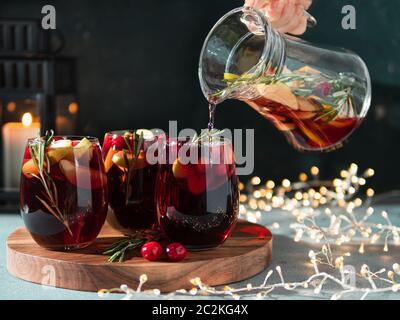 La main femelle verse la sangria d'hiver dans des verres avec tranche de fruits, canneberge et romarin. Arrière-plan sombre de noël avec bougie, décoration Lightti Banque D'Images