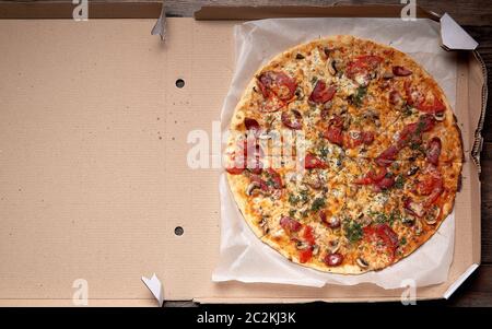 Ronde au four pizza avec saucisses fumées, champignons, tomates, fromage et de l'aneth dans une boîte en carton sur une table en bois, de l'alimentation est coupée en portions, t Banque D'Images