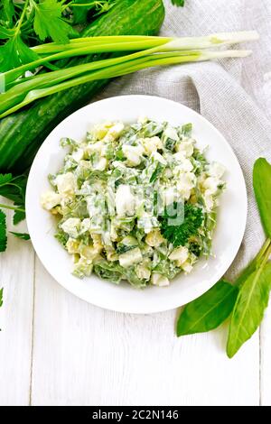 Salade de concombre, d'oseille, de pommes de terre bouillies, d'œufs et d'herbes, garnie de mayonnaise dans une assiette blanche, de persil, d'oignons verts et de serviette de cuisine à l'arrière Banque D'Images