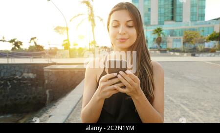 Jeune femme souriante envoyant un message avec smartphone au coucher du soleil avec station balnéaire sur l'arrière-plan. Positive tendance fille blogging dans le réseau social nous Banque D'Images