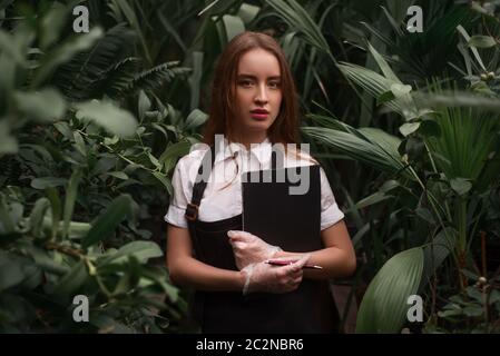 Le jeune fleuriste en tablier tient le presse-papiers dans les mains contre la serre avec des plantes exotiques. Banque D'Images