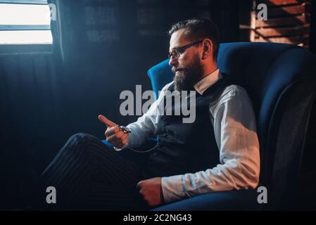 Portrait d'homme barbu à lunettes assis dans un fauteuil, regardant la montre de poche. Écrivain, journaliste, auteur de la littérature, blogger ou poète concept Banque D'Images