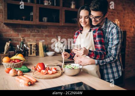 L'amour heureux couple prépare un dîner romantique sur la cuisine. Smiling mari épouse sa femme en tablier qui est en train de préparer une salade de légumes Banque D'Images