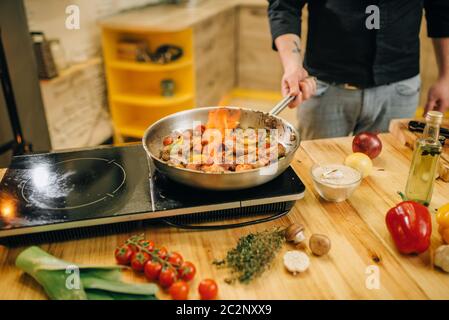 Chef cuisinier masculin cuisant de la viande avec des produits à base de vestable au feu, flamme dans la poêle, cuisine sur fond. Homme préparant du bœuf Banque D'Images