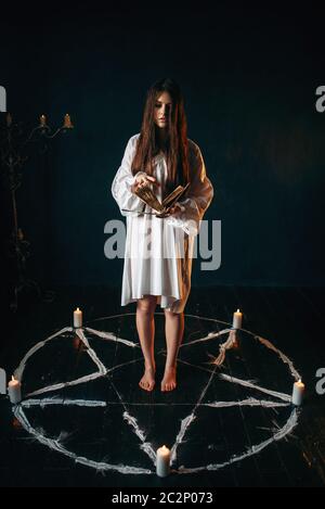 Jeune femme en chemise blanche au centre de pentagram cercle avec bougies et lit un sort, plancher en bois noir. Rituel de magie noire, occulte un Banque D'Images